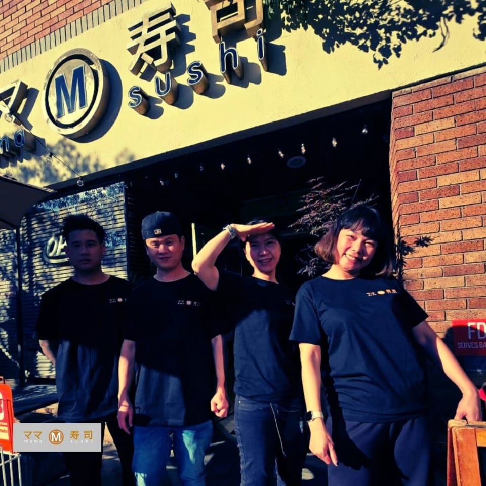 Four Mama M Sushi staff members in black shirts standing outside the restaurant entrance.