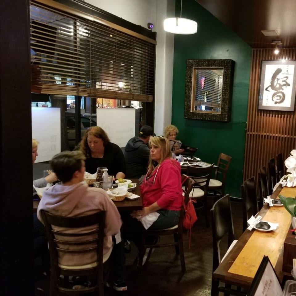 Diners seated at tables and a sushi bar in Mama M Sushi, with green walls and wooden blinds.