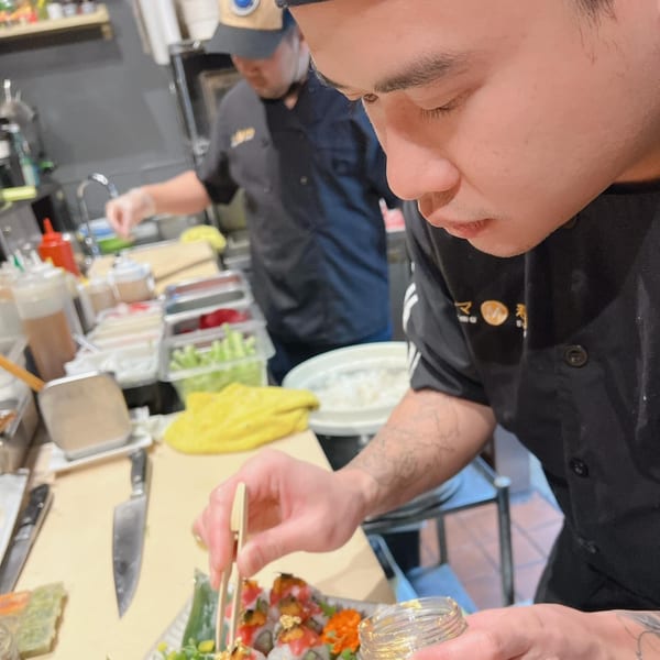 A Mama M Sushi chef meticulously garnishes a sushi roll with gold flakes using tweezers.