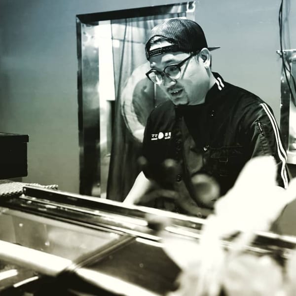 A Mama M Sushi chef in a black uniform and cap preparing sushi behind a counter.