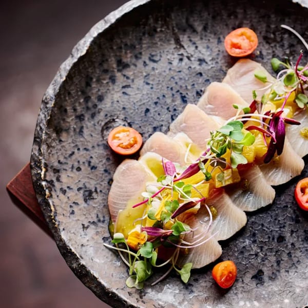 Mama M Sushi's delicate white fish sashimi with citrus, microgreens, and cherry tomatoes on a dark plate.